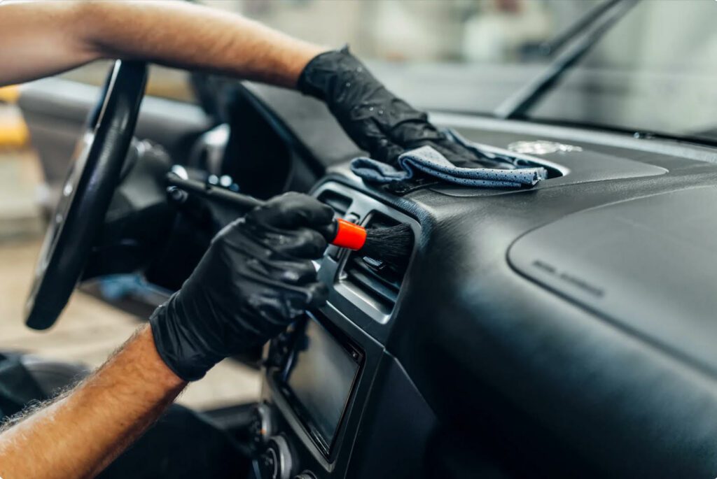 Une personne nettoie l'intérieur d'une voiture avec une brosse et un chiffon, portant des gants noirs.