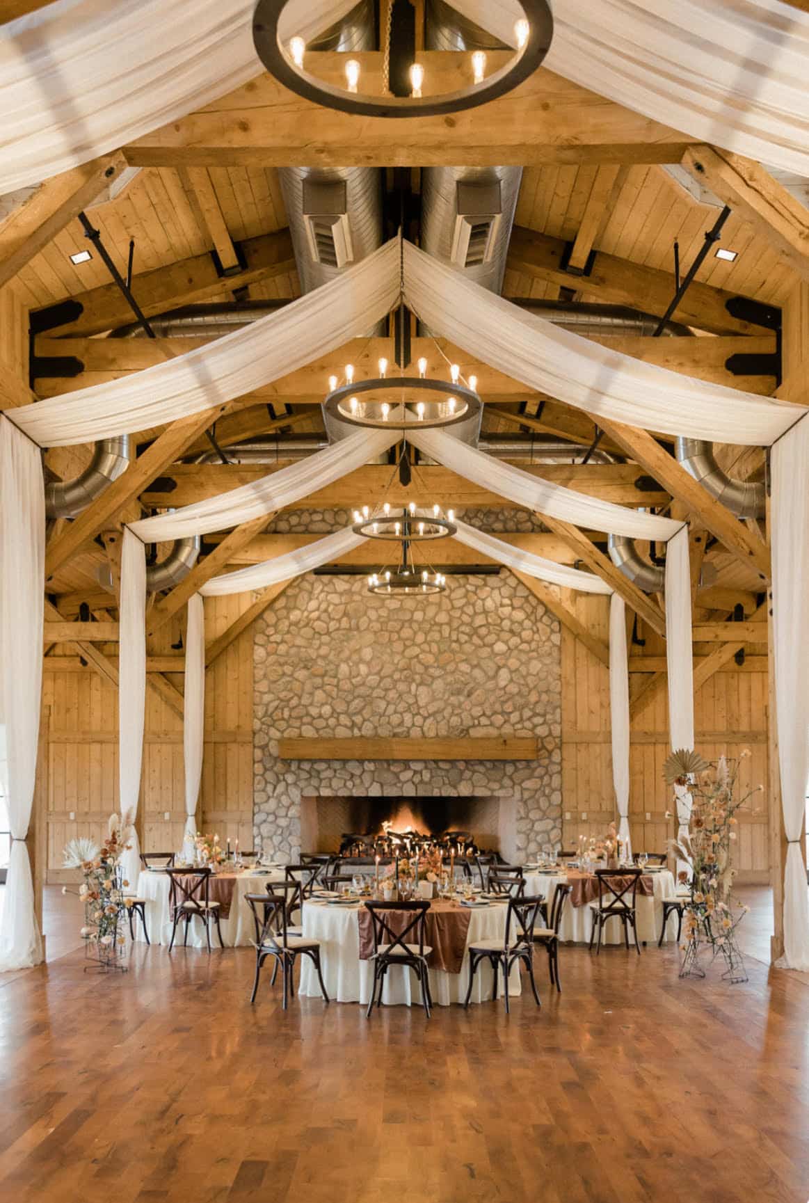 Une salle de réception élégamment décorée avec un toit en bois et des chandeliers suspendus, des tables rondes ornées de nappes blanches et brunes, et une grande cheminée en pierre.